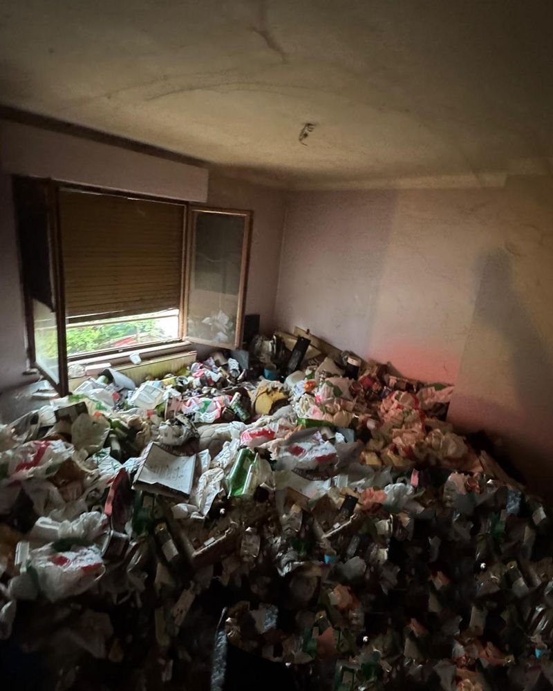 Chambre avant intervention : sacs, bouteilles et déchets accumulés sur tout le sol, jusqu'au rebord de la fenêtre